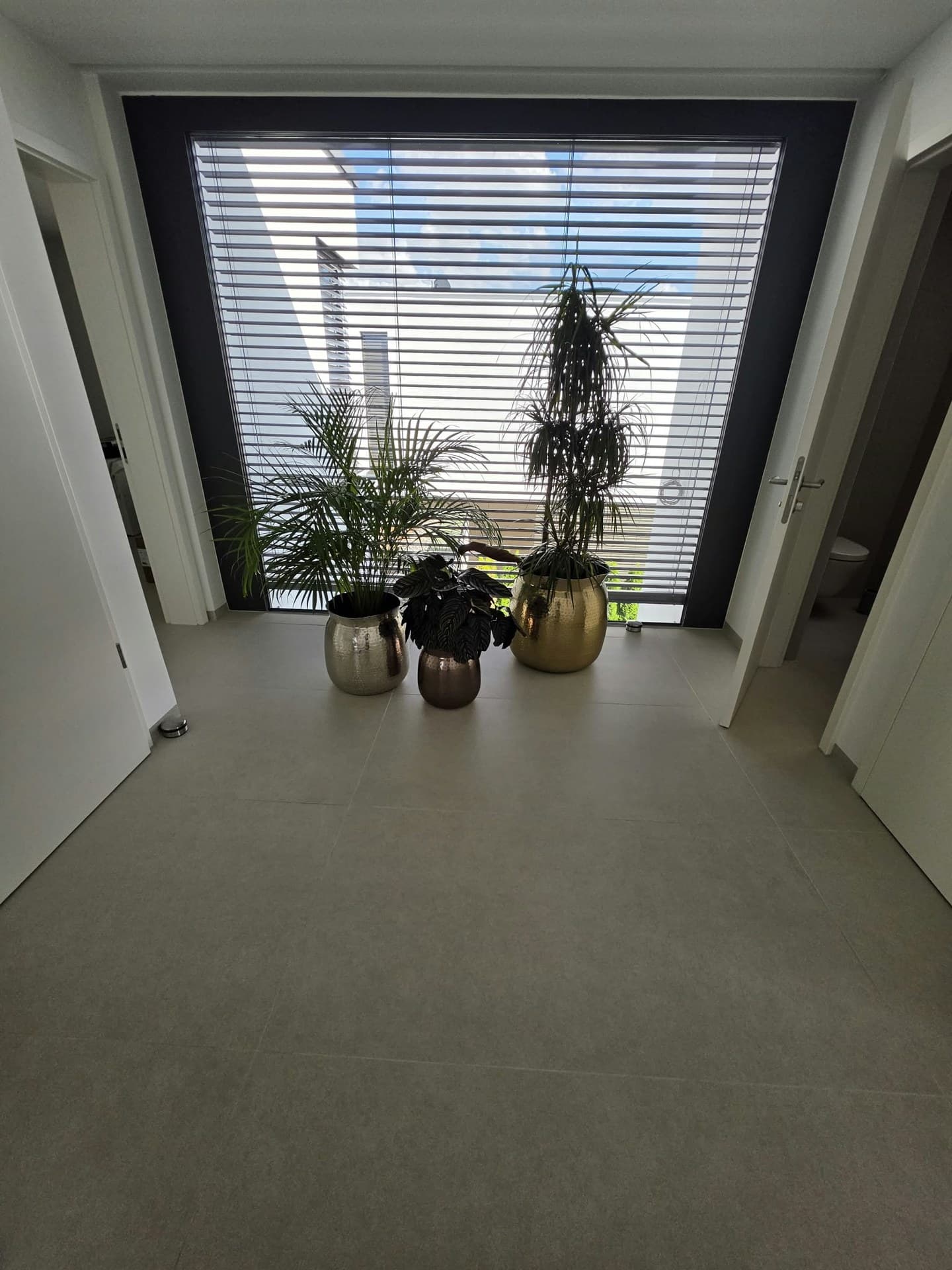 Flur mit großformatigen hellgrauen Bodenfliesen bis zu einem großen Fenster mit Pflanzen in Metalltöpfen